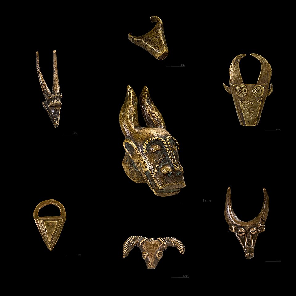 Cast brass Akan gold weight shaped as a bovid head, its horns curving back over a ridged muzzle, photographed against a plain ground.