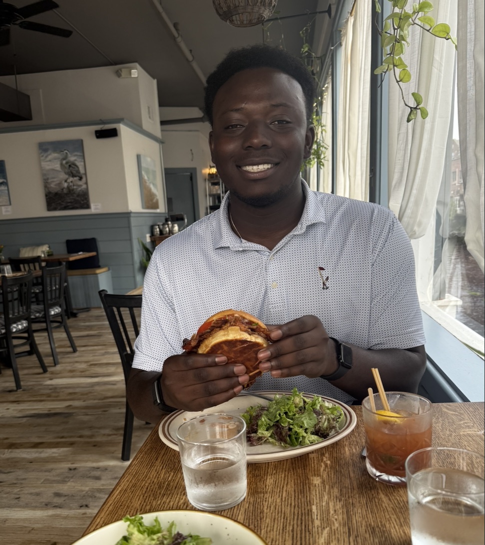 Sitting at a restaurant table by the window, holding a burger with both hands over a plate of salad.