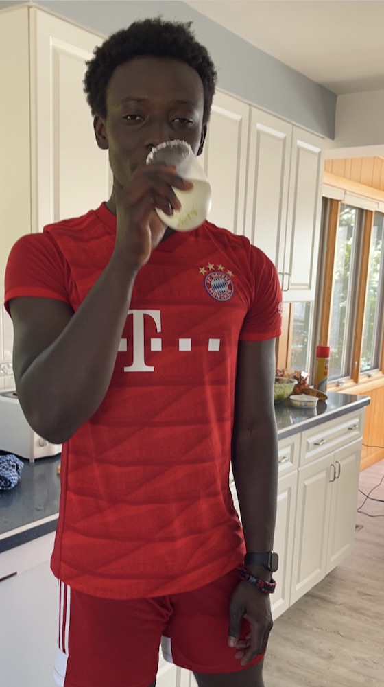Standing in a kitchen drinking a glass of milk, wearing a red Bayern Munich kit.