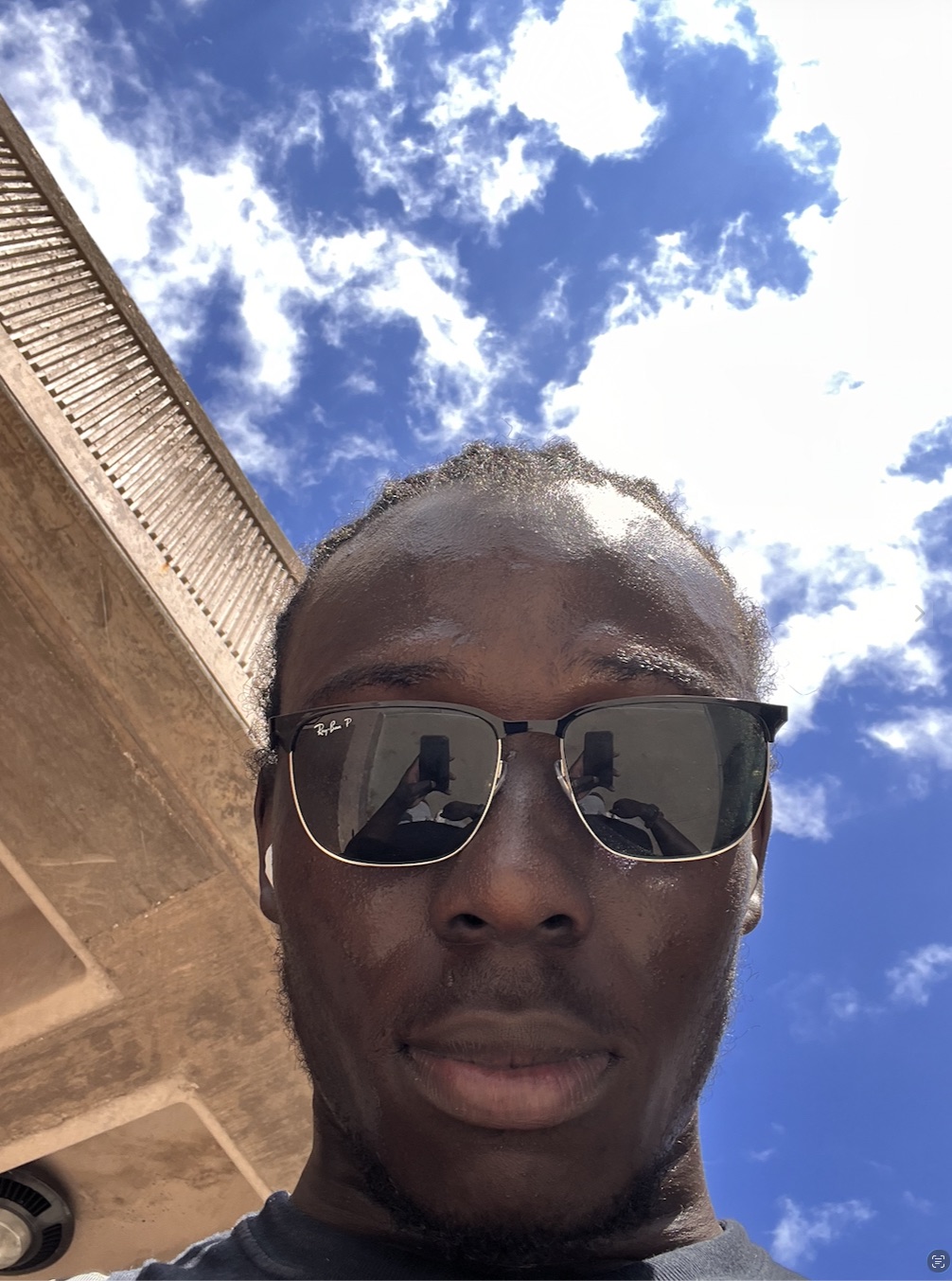 Selfie in Ray-Bans under a concrete stair, cumulus clouds filling the rest of the frame.