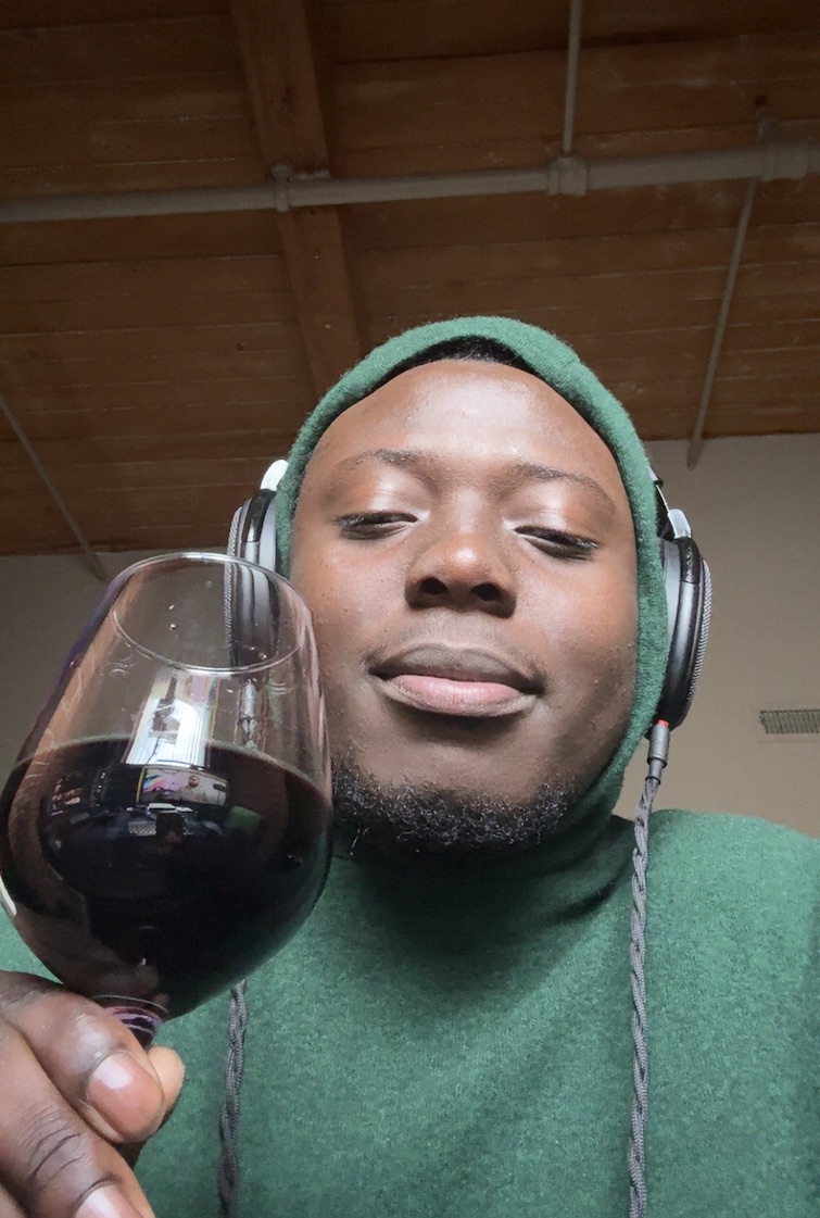 Selfie in a green hooded sweatshirt and wired over-ear headphones, holding a glass of red wine.